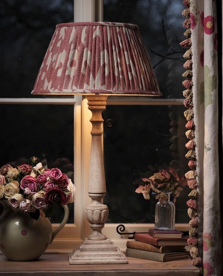 A carved wood table lamp with a pleated floral shade glowing beside a window, fresh roses and a small stack of books