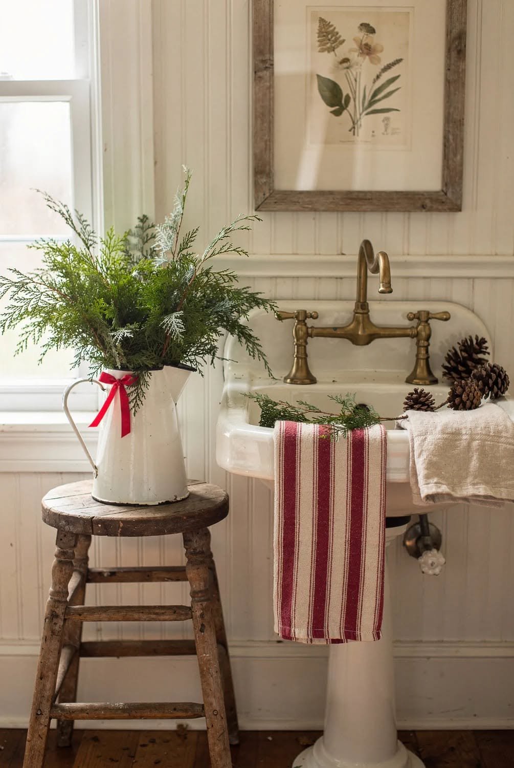 Vintage white pitcher filled with greenery on a wooden stool by the sink