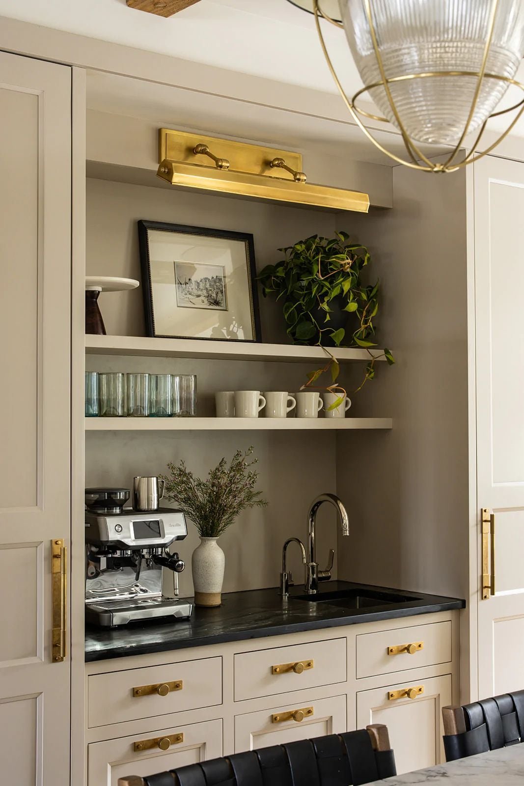 Built-in cabinet nook with an espresso machine, small sink, black counter, brass pulls and a brass picture light