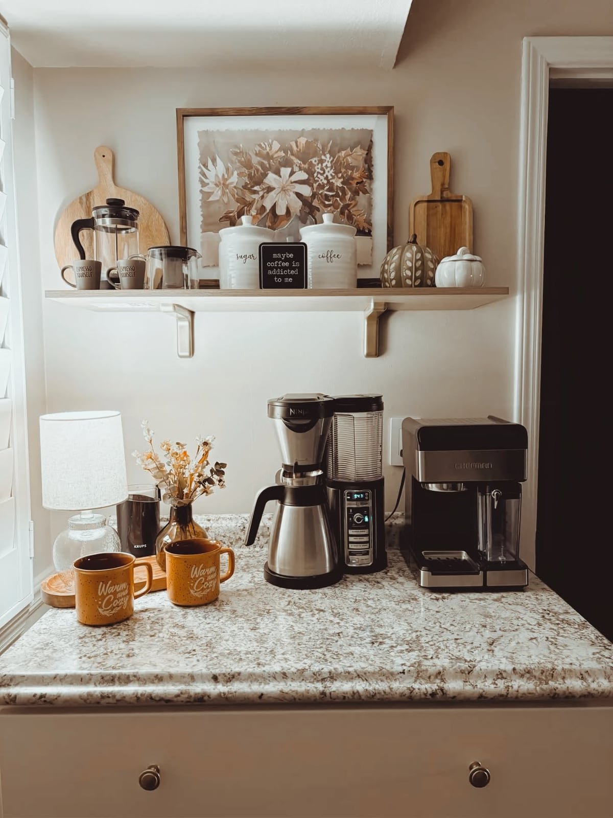 Speckled counter with two coffee makers, two mugs and a small lamp beneath a single floating shelf with framed art and canisters