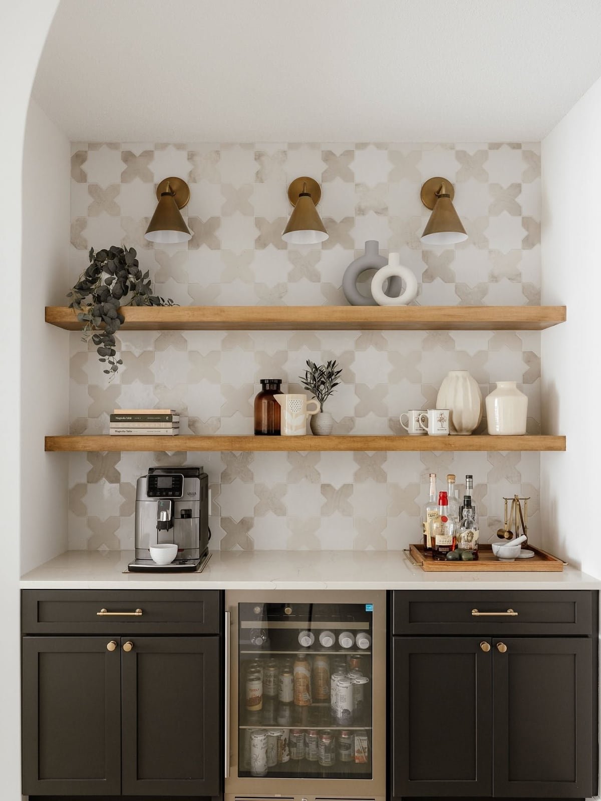 Coffee bar with dark cabinets, a white counter, patterned tile, brass sconces and two wood shelves above a single espresso machine