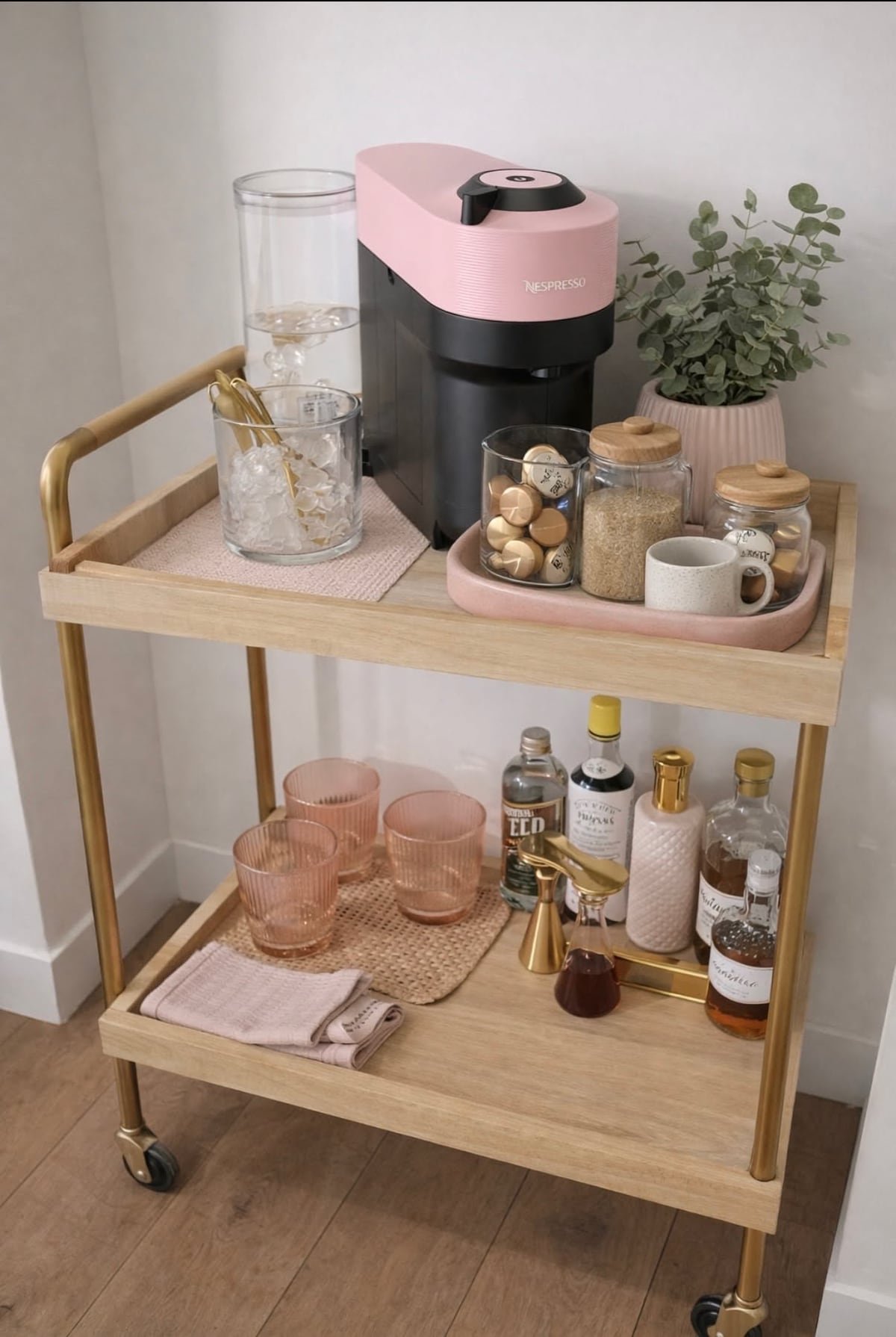 Two-tier wood and brass rolling cart with a pink coffee machine and glass jars on top and pink glasses and bottles on the lower shelf
