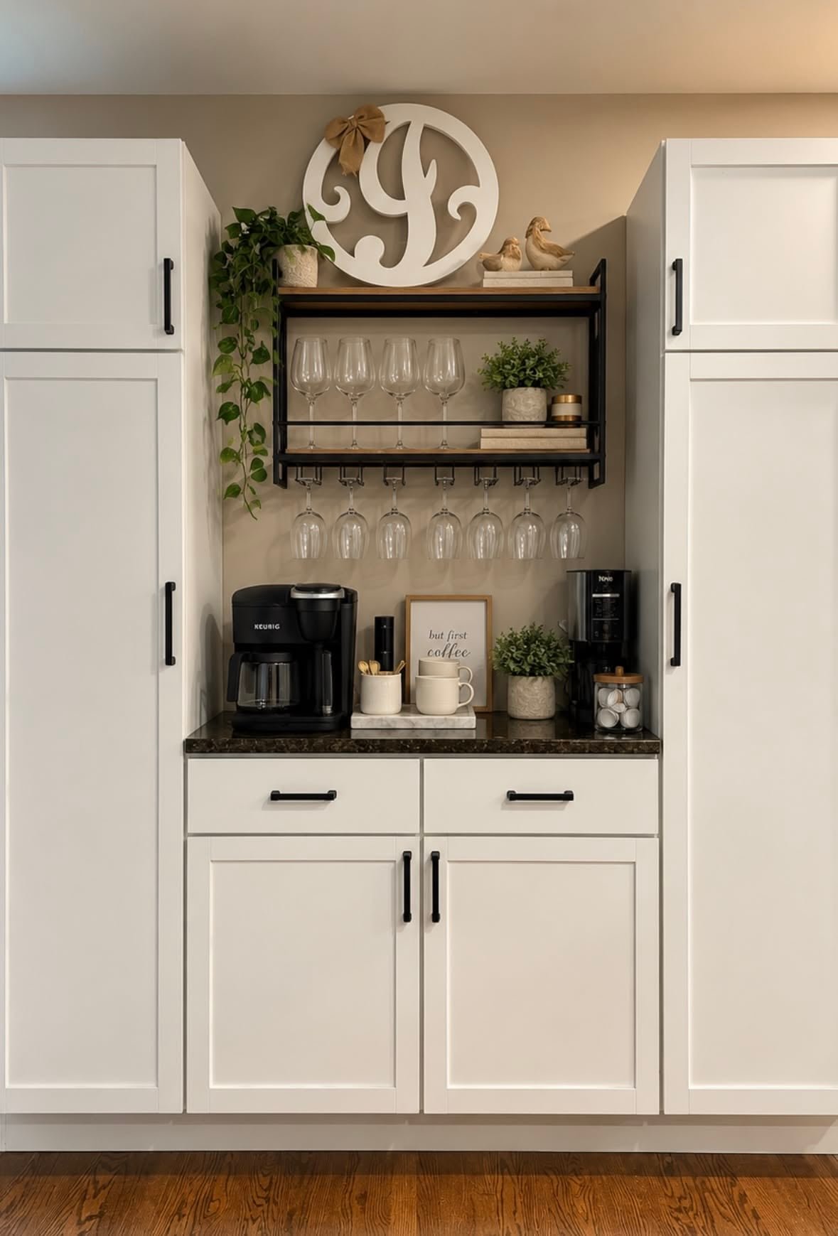 White cabinets with black hardware, a black metal wine glass rack and a coffee maker on a dark counter, with a monogram letter above