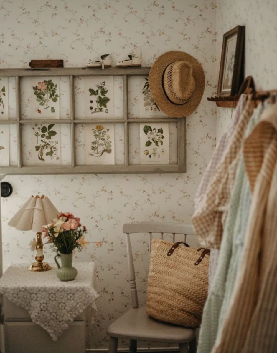 Cottagecore bedroom corner with a straw hat and woven tote hung above a rack of gingham dresses, floral wallpaper and pressed botanical prints