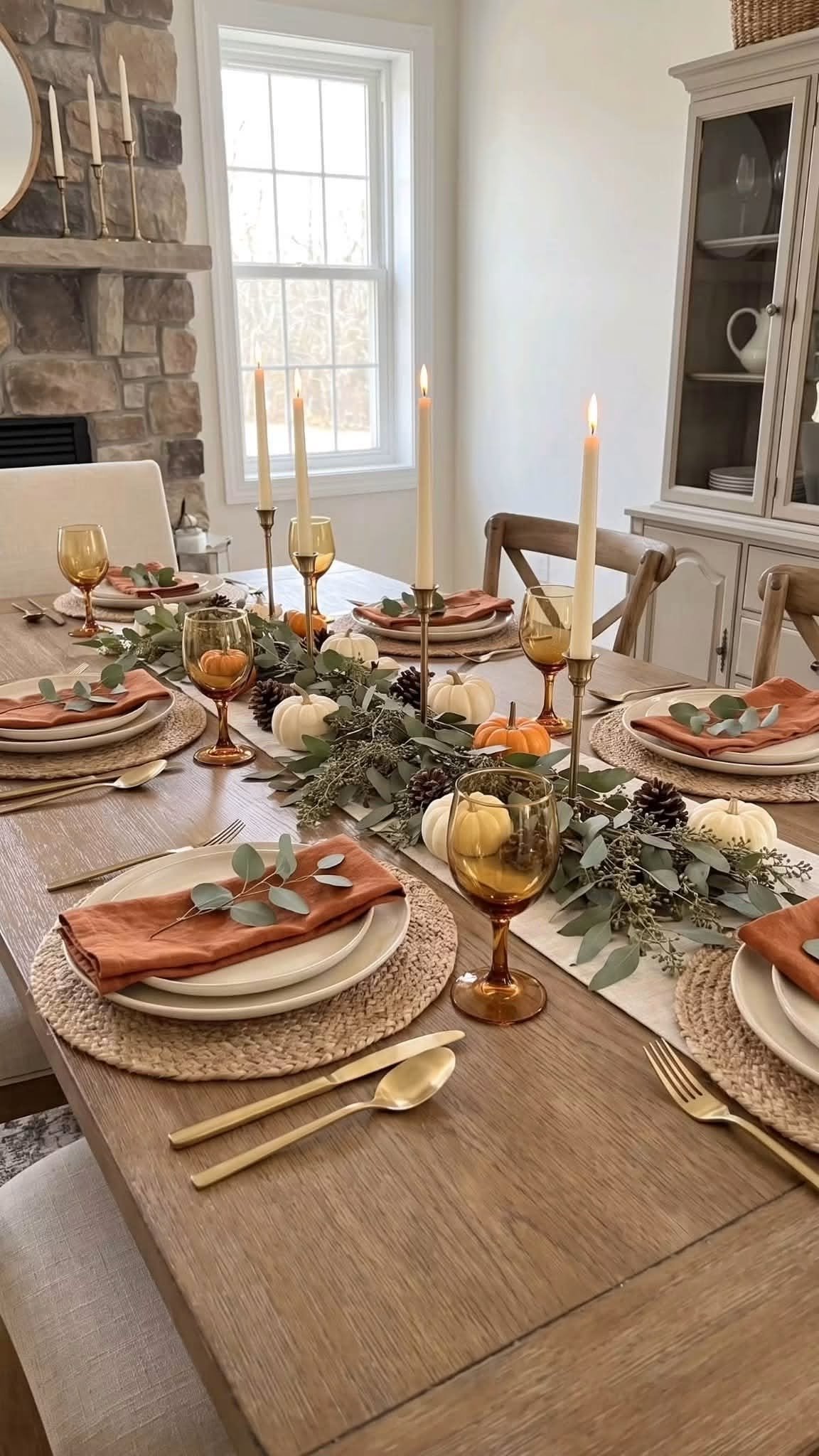 A fall dining table with amber glassware, white pumpkins, and taper candles by a stone fireplace